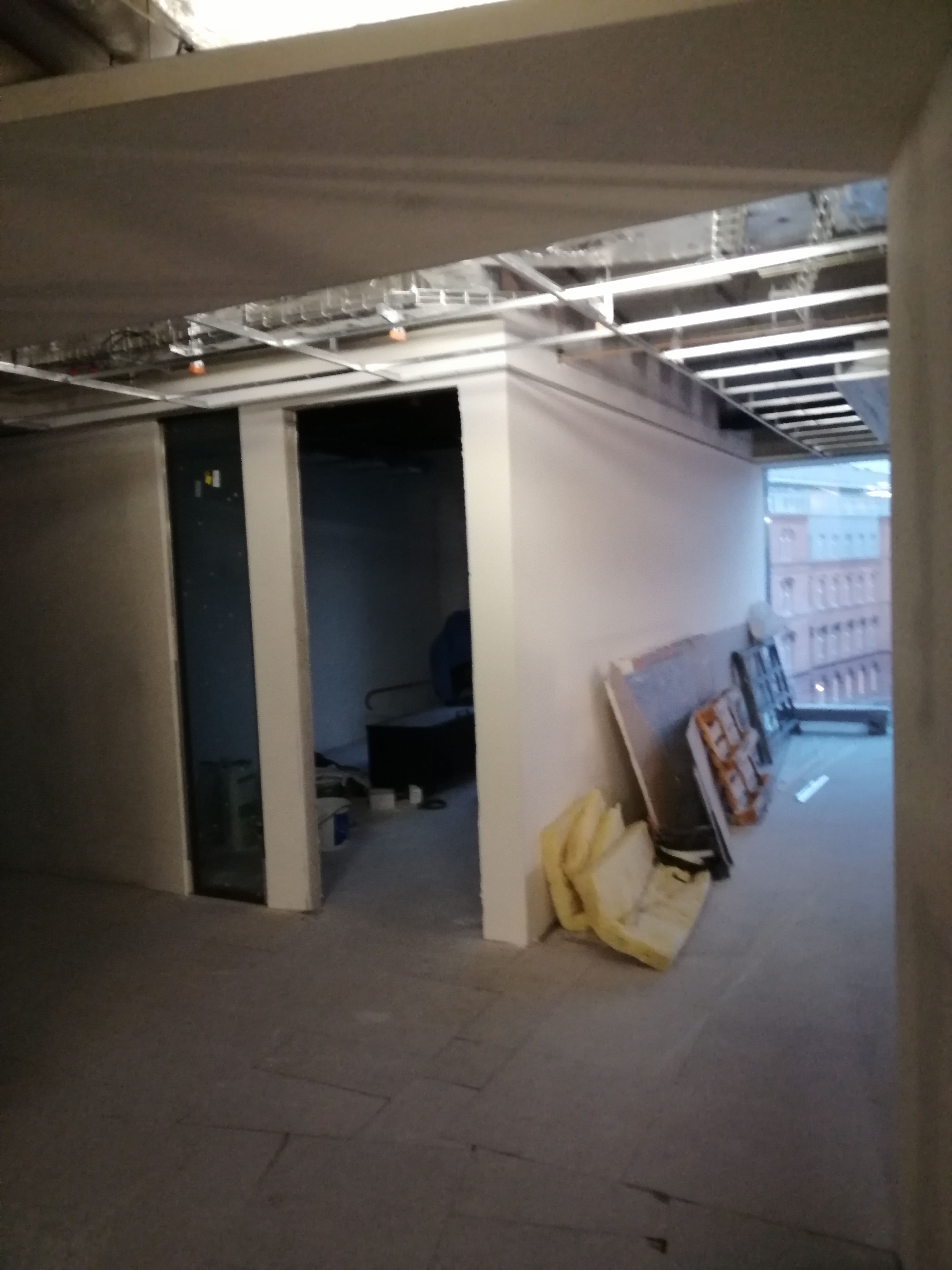 Wnętrze w trakcie prac wykończeniowych, widoczne płyty gipsowe oparte o ścianę, wełna mineralna, surowy sufit z instalacjami i widok na budynek przez okno.