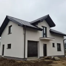 Nowoczesny, dwukondygnacyjny dom z białym tynkiem i ciemną dachówką, widoczny z perspektywy gruntu, z niezagospodarowanym terenem wokół.