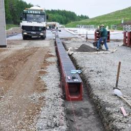 AS Piaseczno Odwodnienia liniowe. - Montaż prefabrykowanego koryta odwadniającego na placu budowy, widoczny pracownik z taczką i ciężarówka firmy Liesnig w tle.