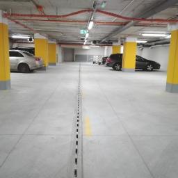 AS Piaseczno Odwodnienia liniowe. - Wnętrze parkingu podziemnego z szarym betonem, żółtymi kolumnami i drenażem liniowym pośrodku, oświetlone lampami sufitowymi.
