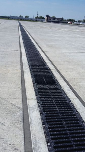 Długa, czarna, żeliwna kratka ściekowa wbudowana w betonową nawierzchnię lotniska z budynkami w tle.
