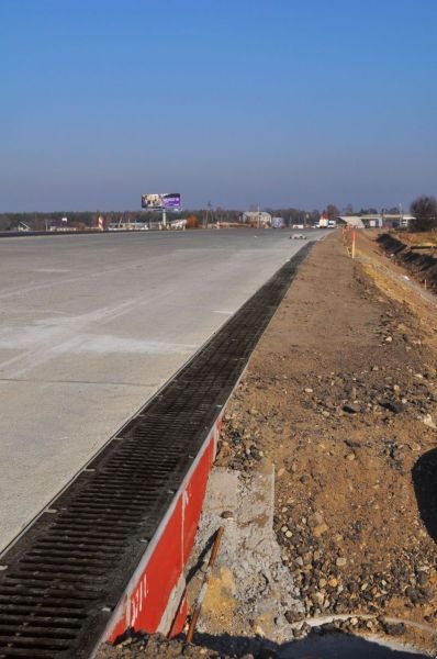 Długi, betonowy plac z zainstalowanym systemem odwadniającym w postaci metalowej kratki; widoczny billboard w tle.