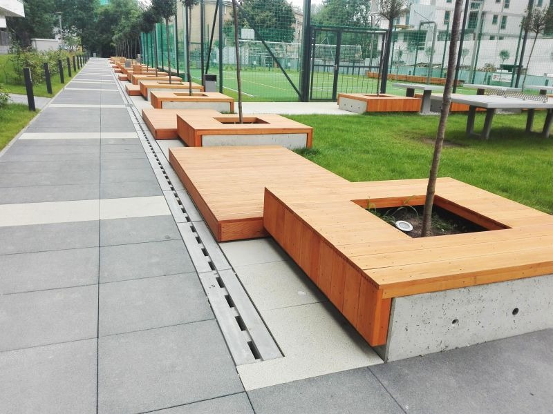 Długa ławka z drewna i betonu z wyciętymi otworami na drzewa, umieszczona wzdłuż chodnika z szarych płyt w parku miejskim. Widoczny system odwadniający wzdłuż ławki.
