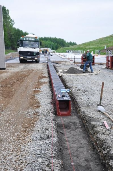 Montaż prefabrykowanego koryta odwadniającego na placu budowy, widoczny pracownik z taczką i ciężarówka firmy Liesnig w tle.