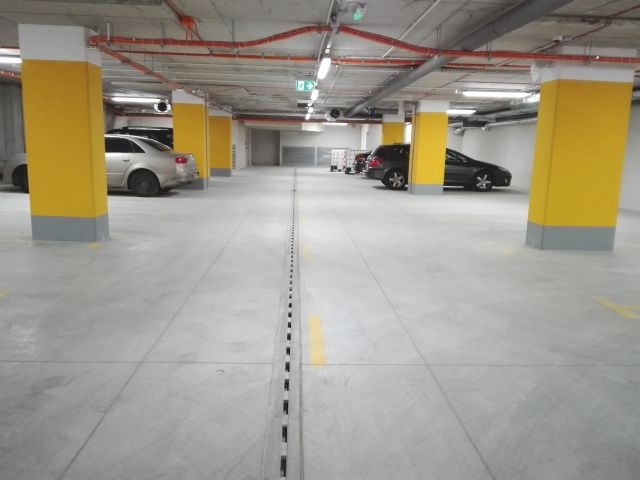 Wnętrze parkingu podziemnego z szarym betonem, żółtymi kolumnami i drenażem liniowym pośrodku, oświetlone lampami sufitowymi.