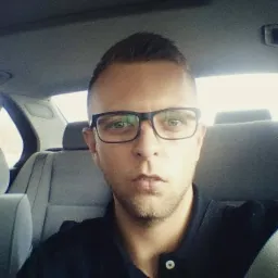 Portret mężczyzny w okularach, siedzącego na tylnym siedzeniu samochodu z szarą tapicerką.