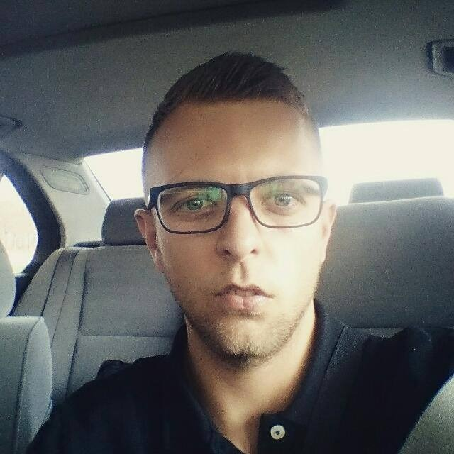 Portret mężczyzny w okularach, siedzącego na tylnym siedzeniu samochodu z szarą tapicerką.