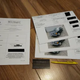 Dokumentacja z opinią rzeczoznawcy motoryzacyjnego, w tym zdjęcia zabytkowego Mercedesa-Benz 126 350SDL z 1991 roku, wizytówka firmy MotoEkspert i żółty długopis na drewnianym podłożu.