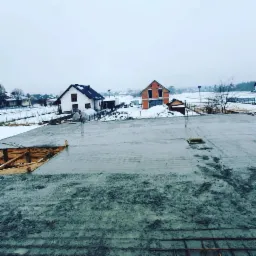 Świeżo wylany, mokry beton na fundamencie domu w zimowej scenerii, widoczne zbrojenia i szalunki, w tle domy w trakcie budowy i śnieżny krajobraz.