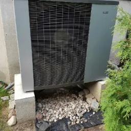 Szara pompa ciepła zamontowana na betonowych blokach przy ścianie budynku, z drobnymi kamieniami na czarnej folii pod spodem i zielonym krzewem obok.