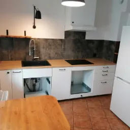 Kuchnia w trakcie montażu: białe szafki z drewnianym blatem, czarny zlew z baterią, płyta indukcyjna, biała lodówka i terakota na podłodze. Jedna szafka otwarta, ukazująca instalację hydrauliczną.