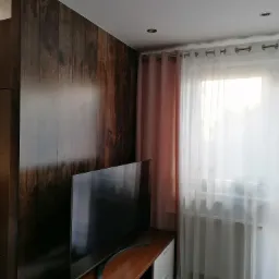 Drewnopodobna ściana działowa z telewizorem w nowoczesnym salonie, widoczne delikatne, różowe zasłony w oknie.