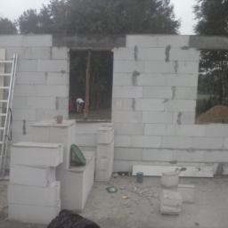 Solidbud - Wznoszenie ściany z białych bloczków betonowych, widoczne okno i drzwi w trakcie budowy, drabina oparta o ścianę, kubek na murze, teren budowy.