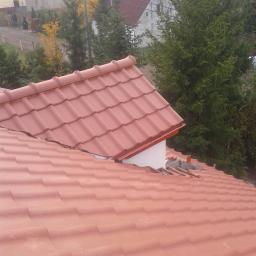 Solidbud - Nowy dach z czerwonej dachówki ceramicznej, widok z góry na fragment połaci z widocznymi szczelinami i fragmentami starego pokrycia, w tle zabudowa jednorodzinna i drzewa.