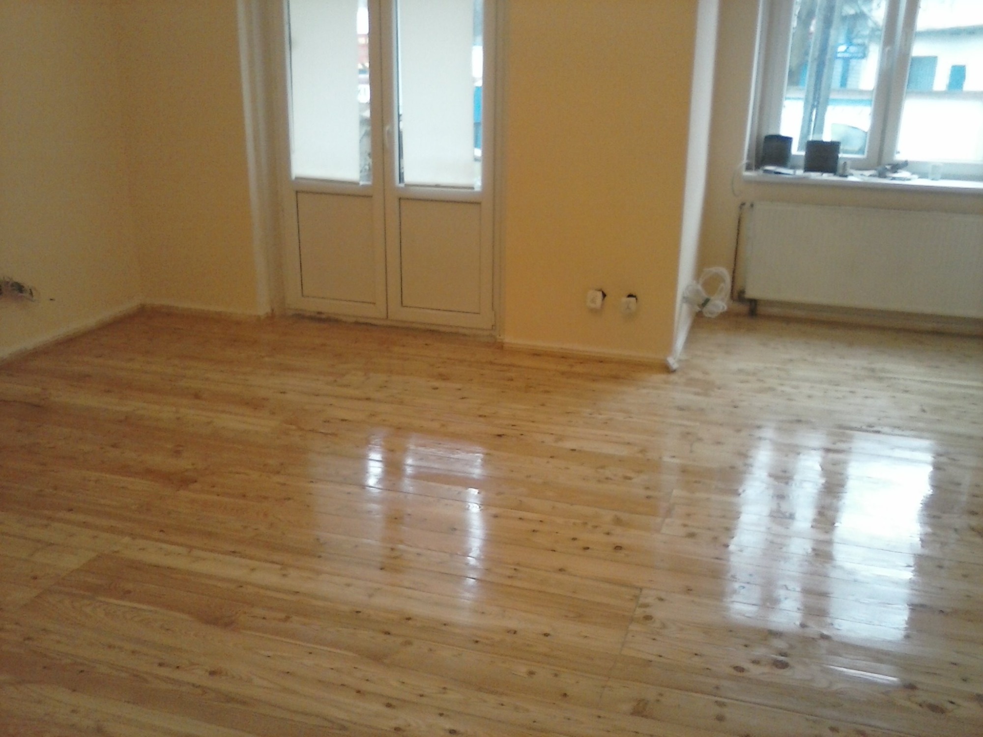 Wycyklinowana, lakierowana podłoga z jasnego drewna w pustym pokoju z białymi drzwiami balkonowymi i grzejnikiem pod oknem.