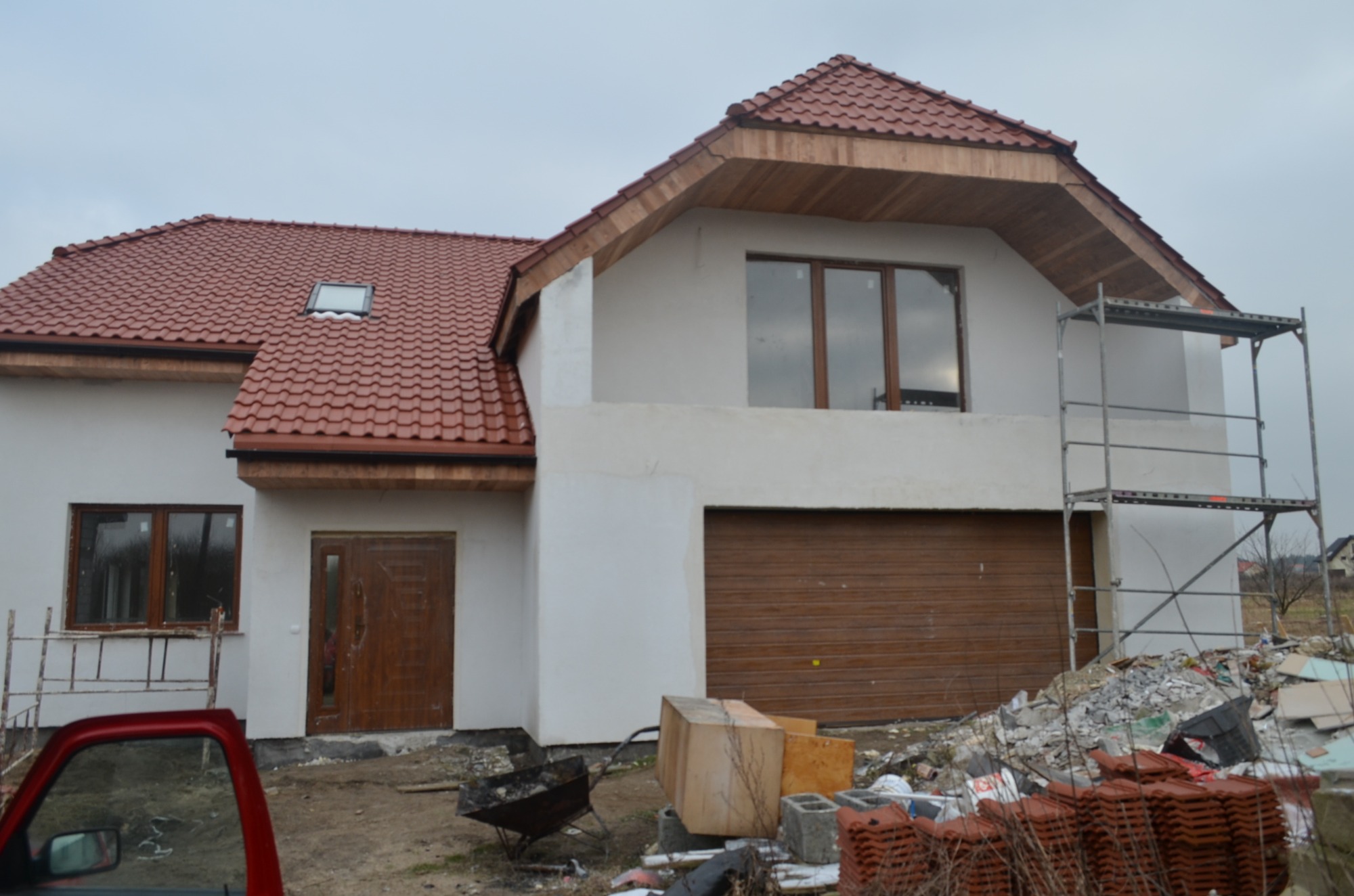 Nowo wybudowany, dwupiętrowy dom z czerwoną dachówką, drewnianą stolarką okienną i bramą garażową w trakcie wykończenia, widoczne rusztowania i materiały budowlane.