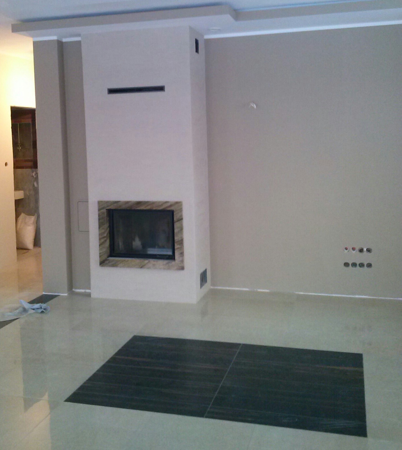 Wykończone wnętrze z kominkiem o obudowie z marmuru, gładkie ściany w odcieniach beżu, błyszcząca podłoga z ciemnymi płytkami i wystającymi gniazdkami elektrycznymi.