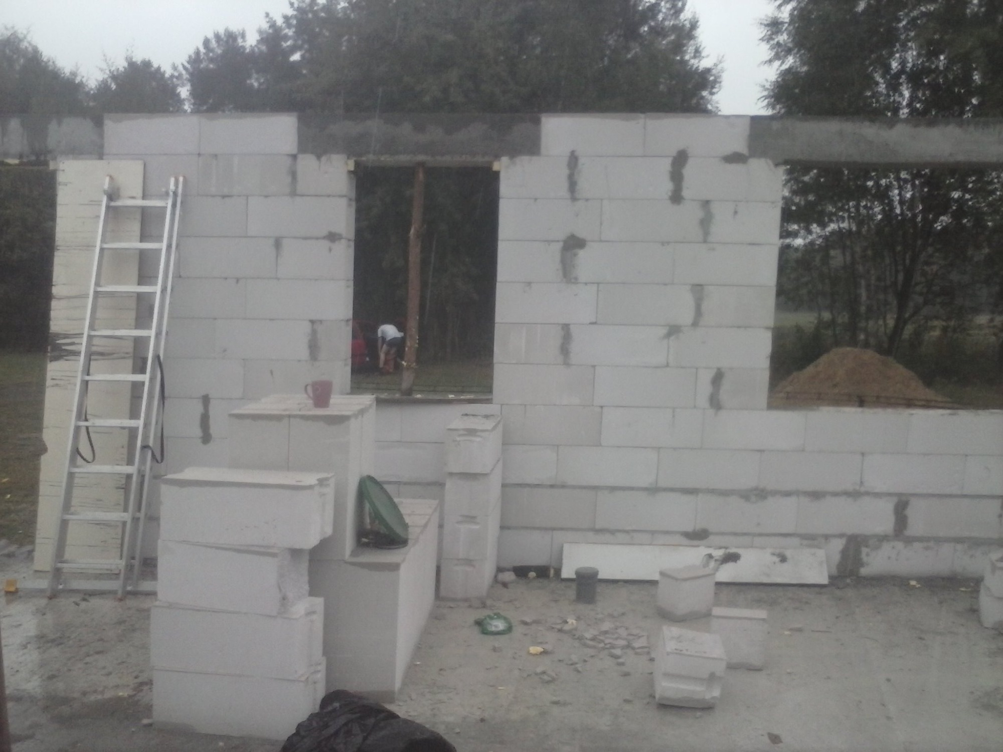 Wznoszenie ściany z białych bloczków betonowych, widoczne okno i drzwi w trakcie budowy, drabina oparta o ścianę, kubek na murze, teren budowy.