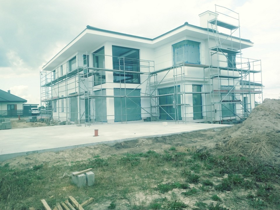 Dwukondygnacyjny, biały dom w trakcie budowy, otoczony rusztowaniami, z zabezpieczonymi folią oknami i widocznym przygotowanym terenem.