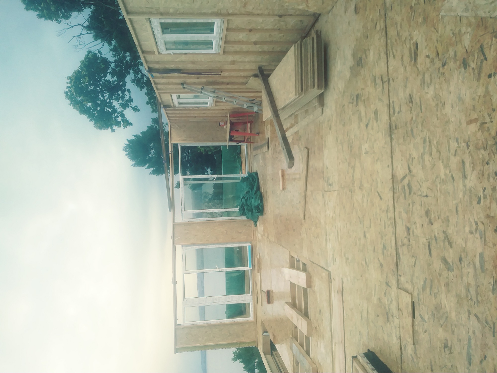 Konstrukcja domu w stanie surowym zamkniętym, ściany obite płytami OSB, widoczne okna PCV i drzwi balkonowe, na podłodze leżą płyty OSB, na ścianie oparta drabina.