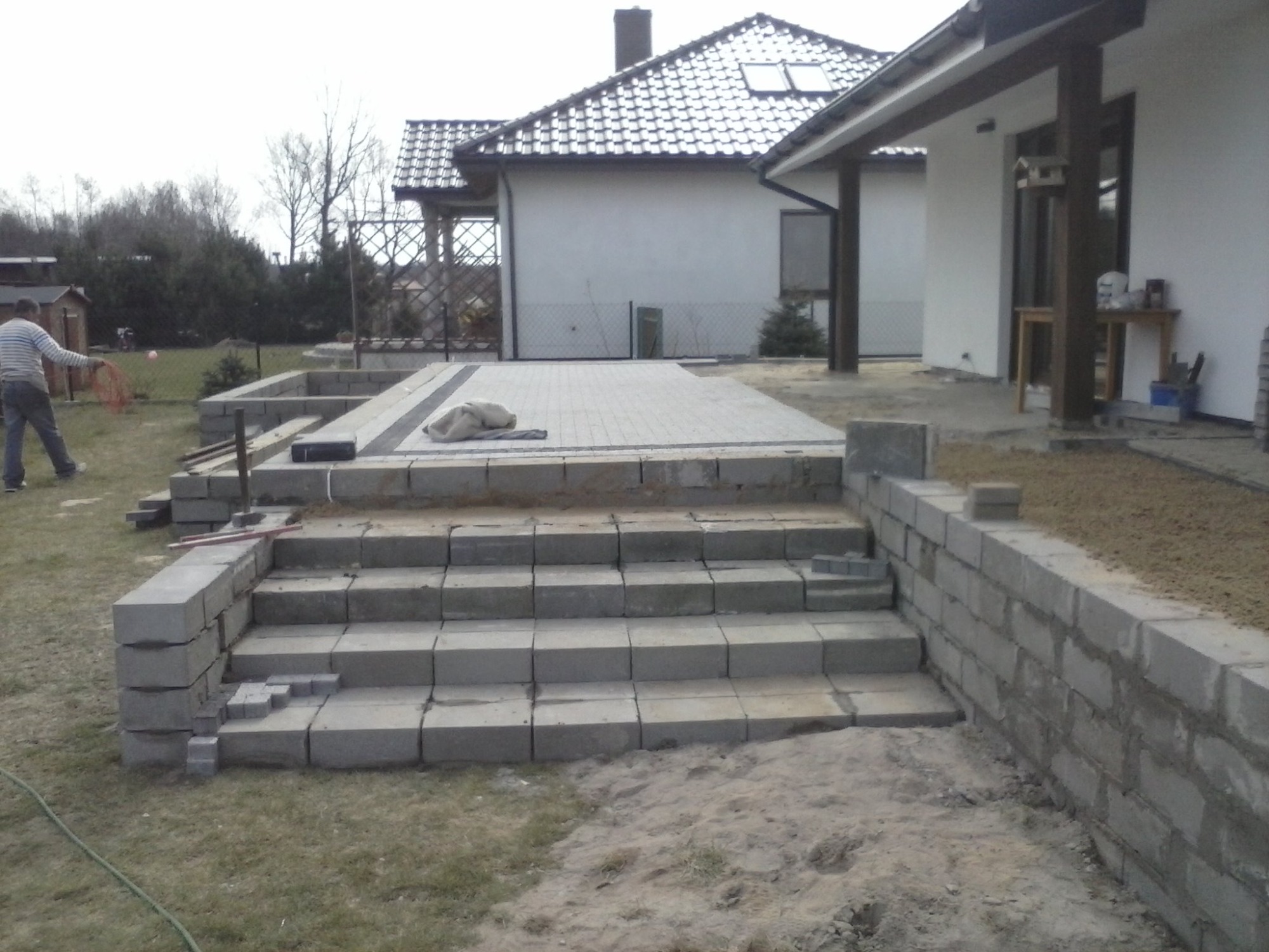 Budowa tarasu z kamiennymi schodami i kostką brukową na posesji z domem jednorodzinnym w tle, widoczny mężczyzna z narzędziami.
