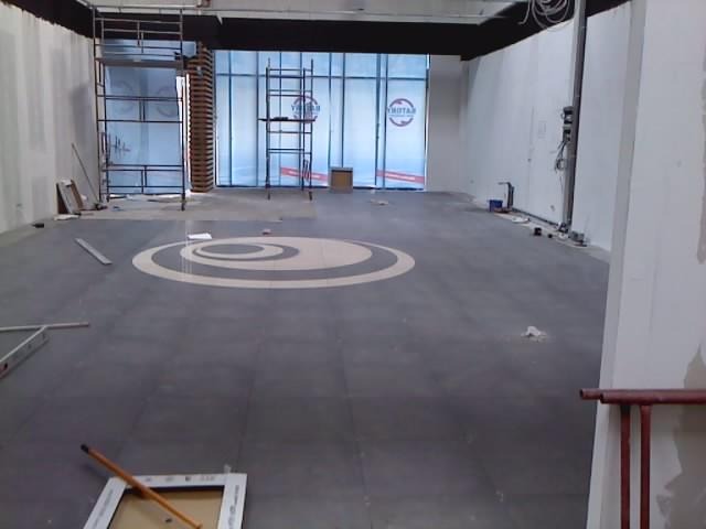 Wnętrze komercyjne w trakcie prac wykończeniowych: szare płytki podłogowe z dekoracyjnym wzorem koła, rusztowania przy oknach i ściany przygotowane do malowania.