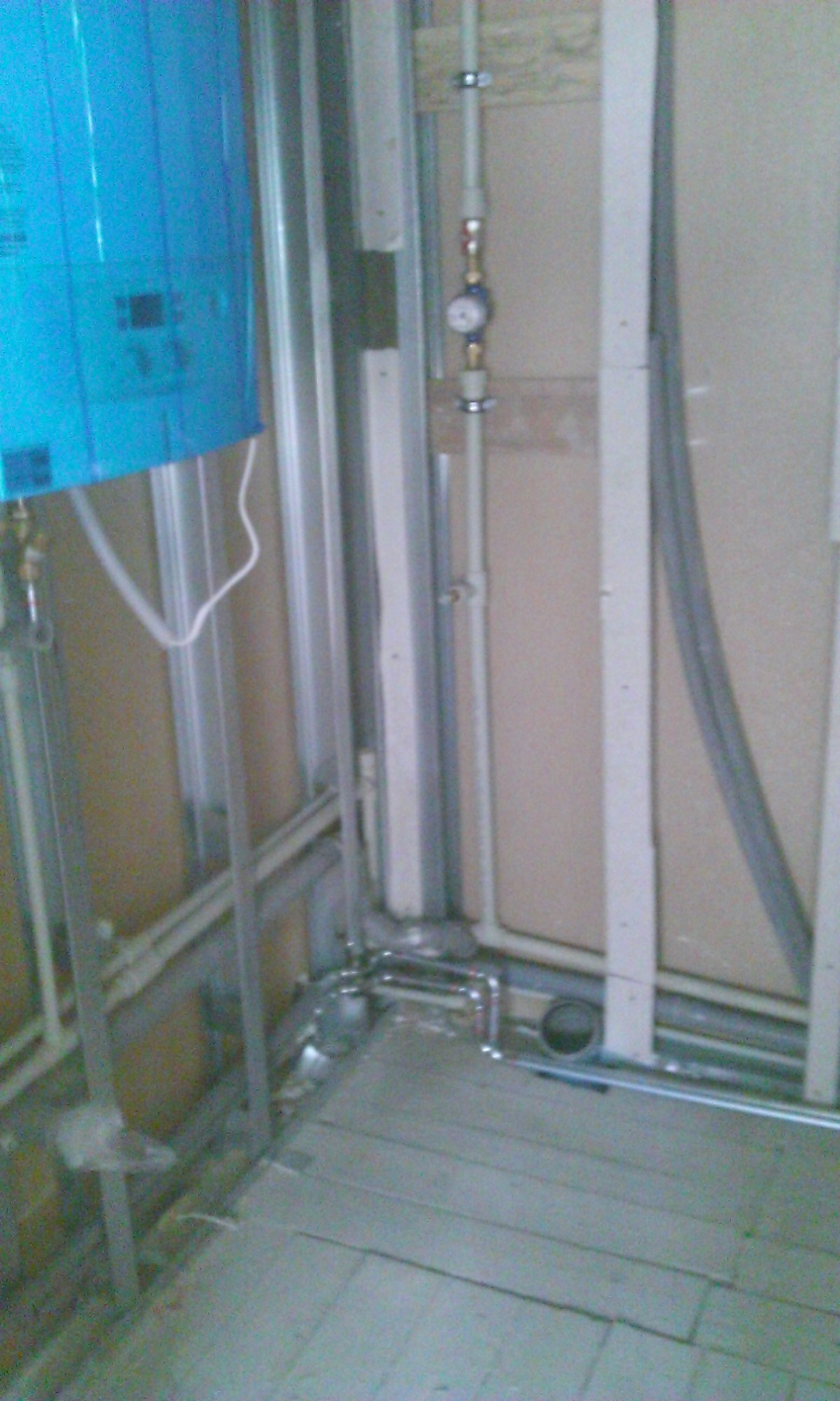 Wnętrze w trakcie prac instalacyjnych: niebieski zasobnik ciepłej wody, metalowa konstrukcja ścianki działowej, rury wodne z manometrem, szara podłoga z płyt.