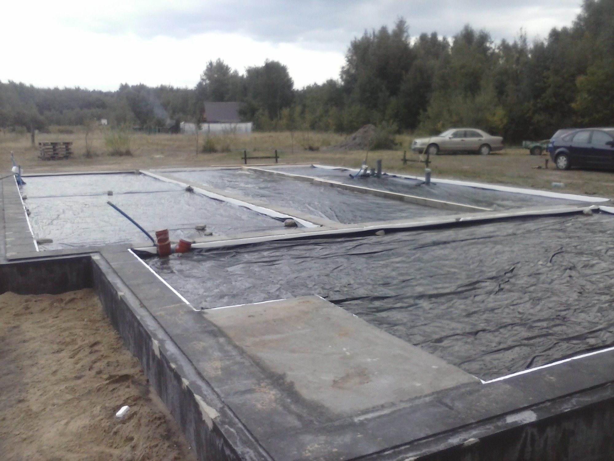Fundament domu w trakcie budowy, zabezpieczony czarną folią, widoczne wystające rury, w tle drzewa i samochody.