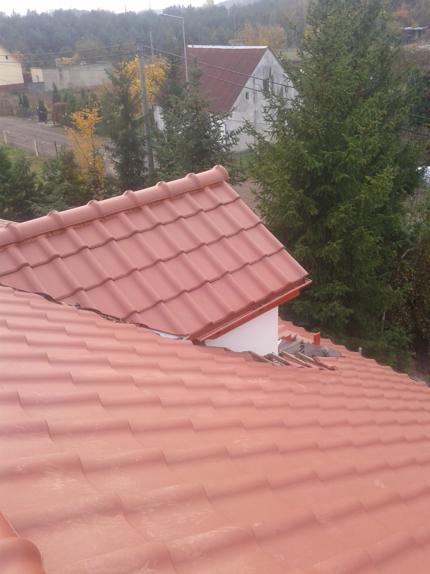 Nowy dach z czerwonej dachówki ceramicznej, widok z góry na fragment połaci z widocznymi szczelinami i fragmentami starego pokrycia, w tle zabudowa jednorodzinna i drzewa.
