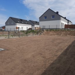 Kamilo Kamil Hubrich - Przygotowany teren pod trawnik lub ogród przed szeregiem nowych domów z szarymi dachami i białymi ścianami, ogrodzony siatką.
