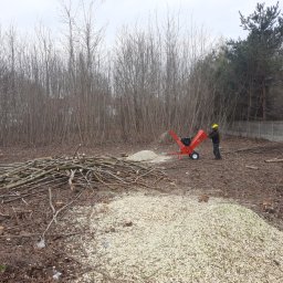 Kamilo Kamil Hubrich - Mężczyzna w żółtym kasku obsługuje czerwoną rębarkę do gałęzi, tworzącą pryzmę zrębków na terenie po wycince drzew, z widocznymi pniami i stertą gałęzi w tle.