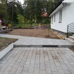 Ułożona kostka brukowa prowadząca do trawnika przed domem z młodymi drzewkami i ozdobnym ażurowym ogrodzeniem.