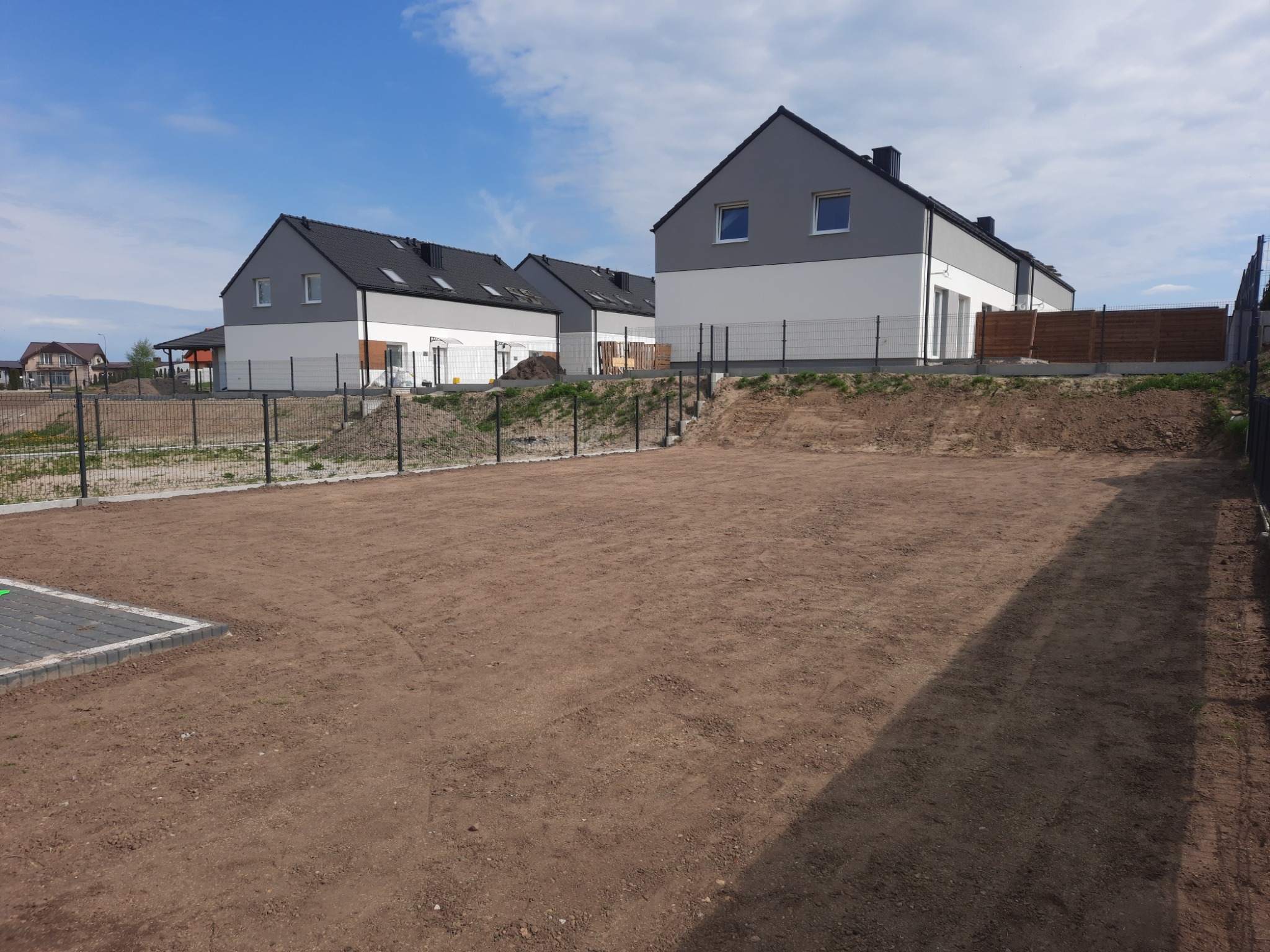 Przygotowany teren pod trawnik lub ogród przed szeregiem nowych domów z szarymi dachami i białymi ścianami, ogrodzony siatką.