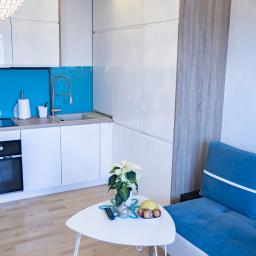 Meble Muzyka - Nowoczesna kuchnia z białymi, lakierowanymi frontami, turkusowym panelem nad blatem i drewnopodobnym wysokim modułem, widziana z perspektywy salonu z niebieską kanapą i białym stolikiem kawowym.