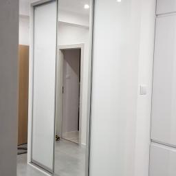 Meble Muzyka - Biała szafa wnękowa z przesuwnymi drzwiami, z jednym lustrzanym panelem, w nowoczesnym wnętrzu z szarą podłogą.