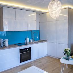 Meble Muzyka - Nowoczesna kuchnia z białymi, lakierowanymi frontami, drewnopodobnym wykończeniem boków i turkusowym panelem szklanym nad blatem, oświetlona wiszącą, kryształową lampą.