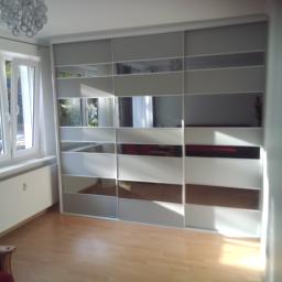 Meble Muzyka - Biała szafa przesuwna z lustrzanymi panelami w pustym pokoju z parkietem, widoczny fragment krzesła z czerwonym obiciem, oświetlenie sufitowe w tle.