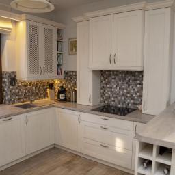 Meble Muzyka - Jasna kuchnia z drewnianymi frontami, mozaiką nad blatem, oświetleniem LED pod szafkami i elementami dekoracyjnymi jak kwiaty i talerze na ścianie.