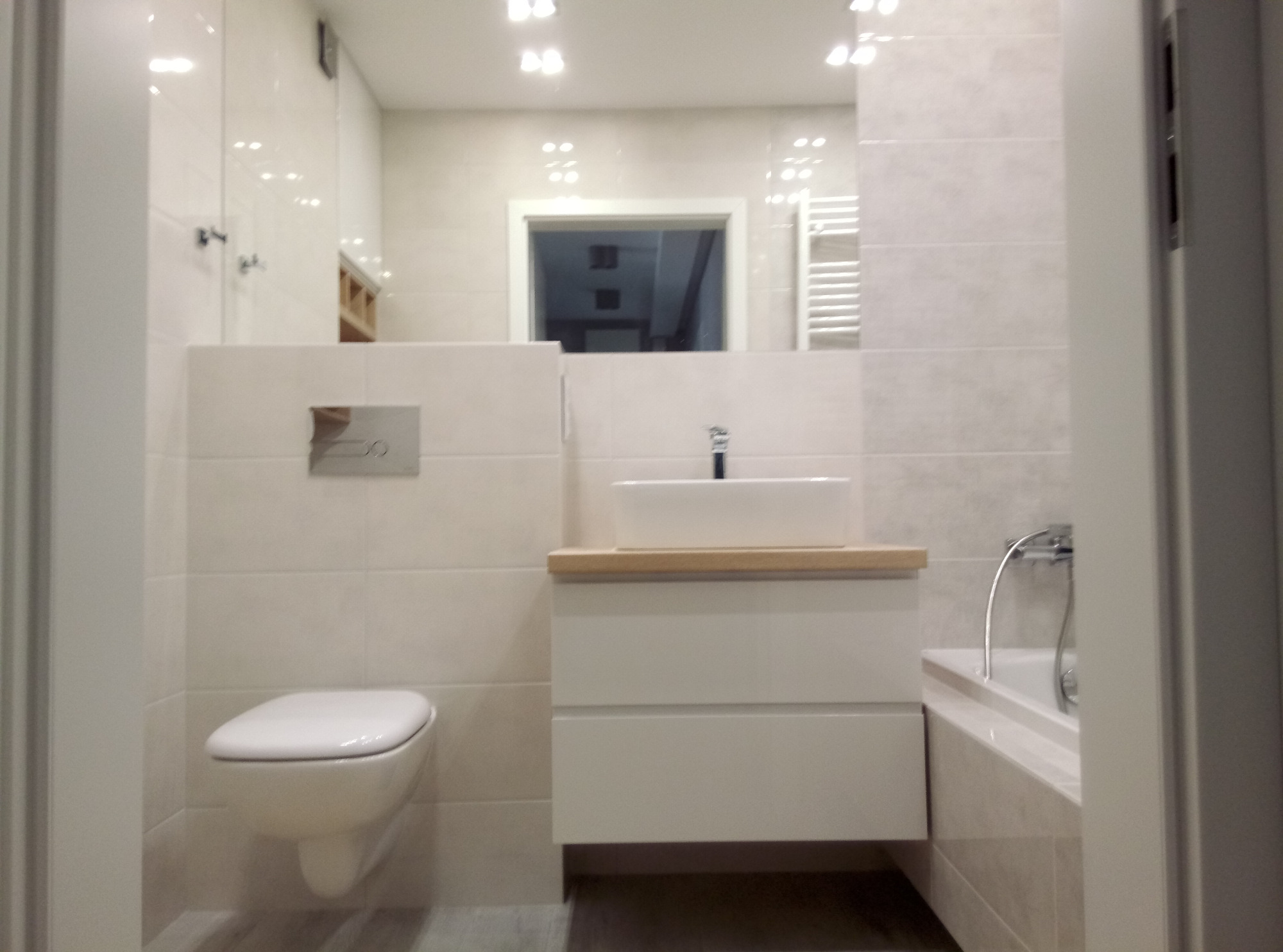 Nowoczesna łazienka w jasnych, kremowych odcieniach z podwieszaną toaletą i umywalką nablatową na szafce z drewnianym blatem, widoczna wanna z chromowaną baterią, odbicie wnętrza w dużym lustrze...