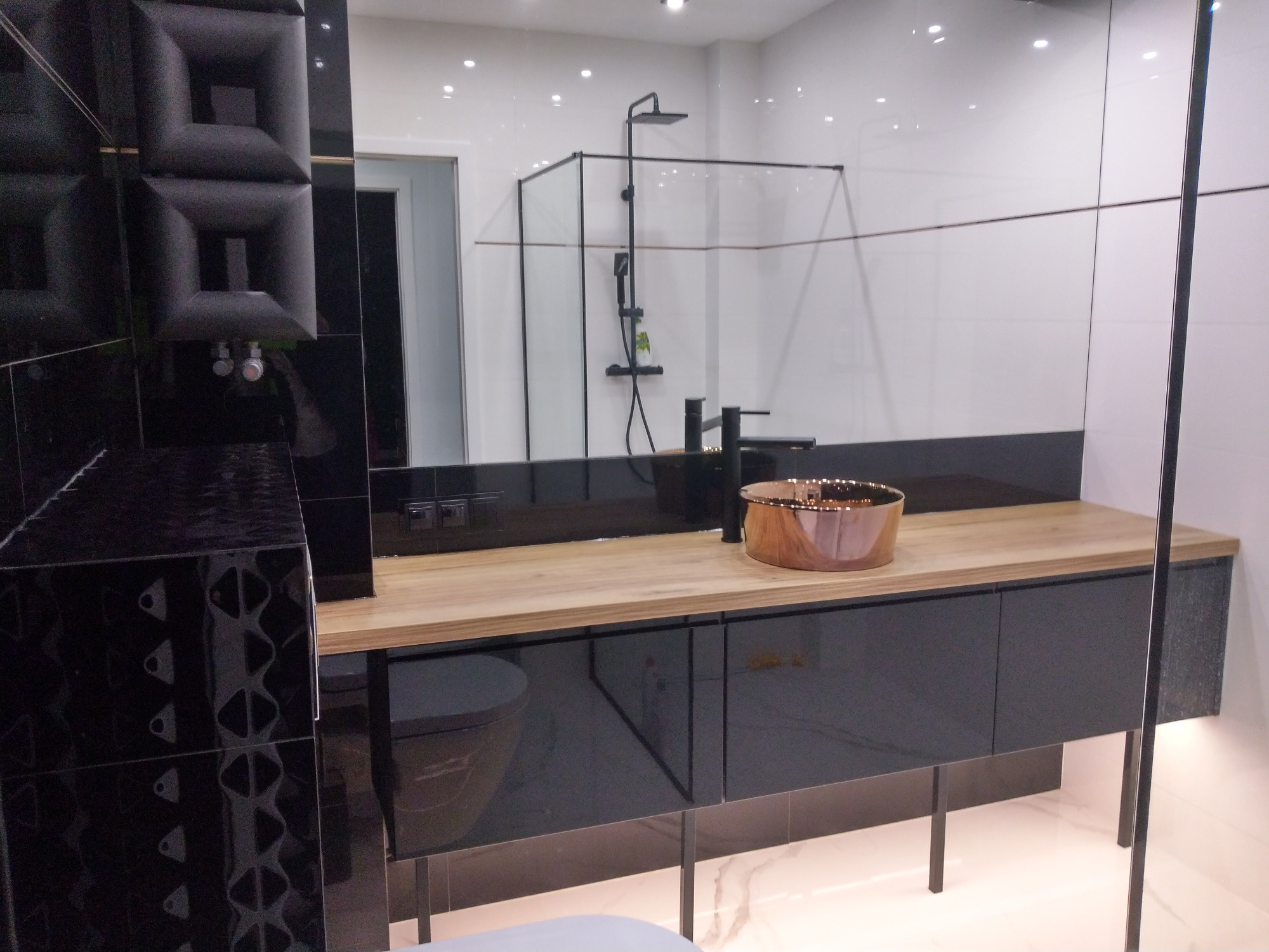 Nowoczesna łazienka z czarnymi szafkami pod umywalką z drewnianym blatem, miedziana umywalka nablatowa, kabina prysznicowa z czarną ramą, czarna armatura i dekoracyjna ściana z czarnych paneli 3D.