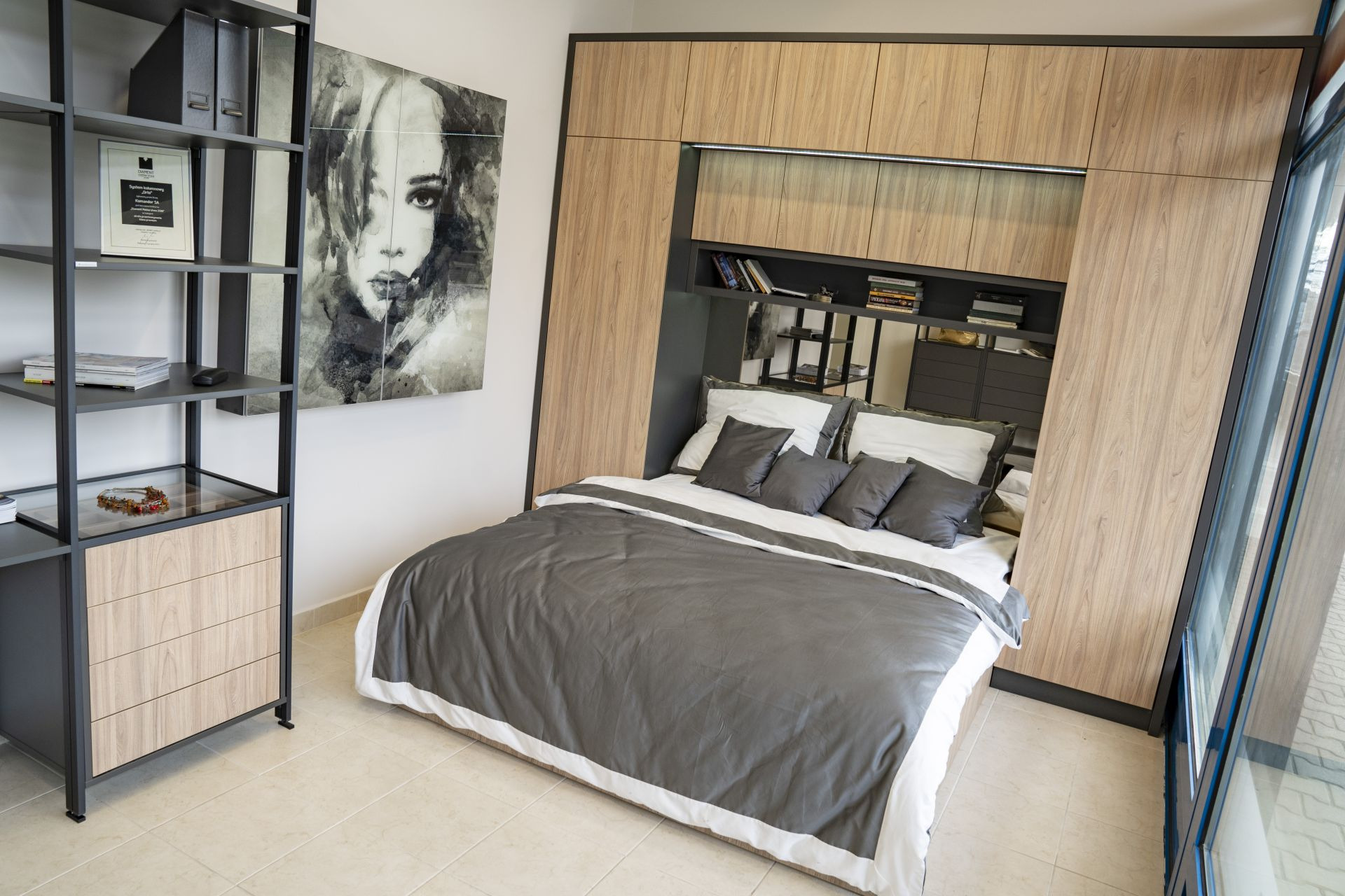 Nowoczesna sypialnia z łóżkiem chowanym w zabudowie z drewnopodobnej płyty, uzupełniona półkami i oświetleniem LED, obok regał z szufladami i dekoracją, na ścianie obraz.