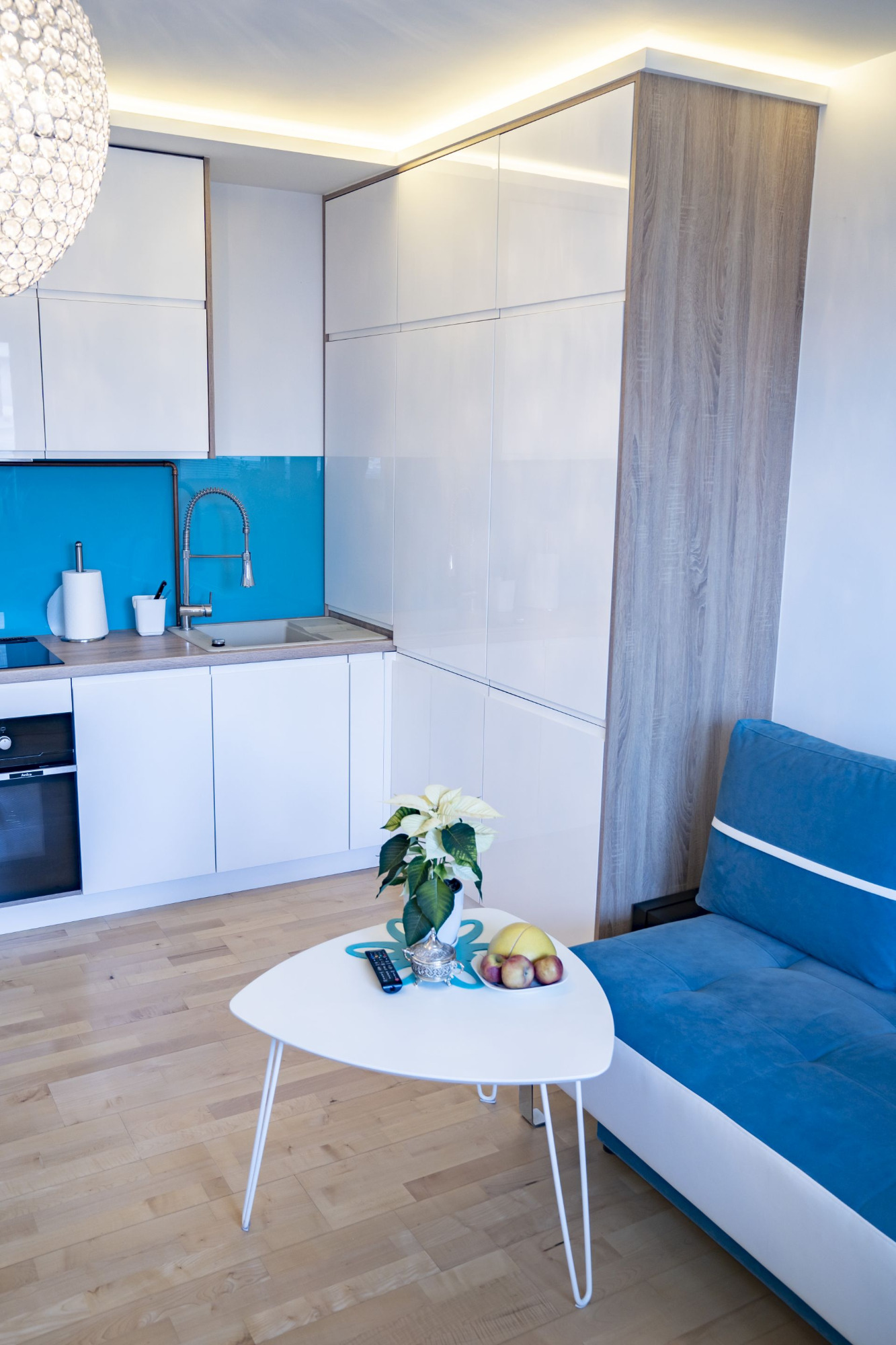 Nowoczesna kuchnia z białymi, lakierowanymi frontami, turkusowym panelem nad blatem i drewnopodobnym wysokim modułem, widziana z perspektywy salonu z niebieską kanapą i białym stolikiem kawowym.