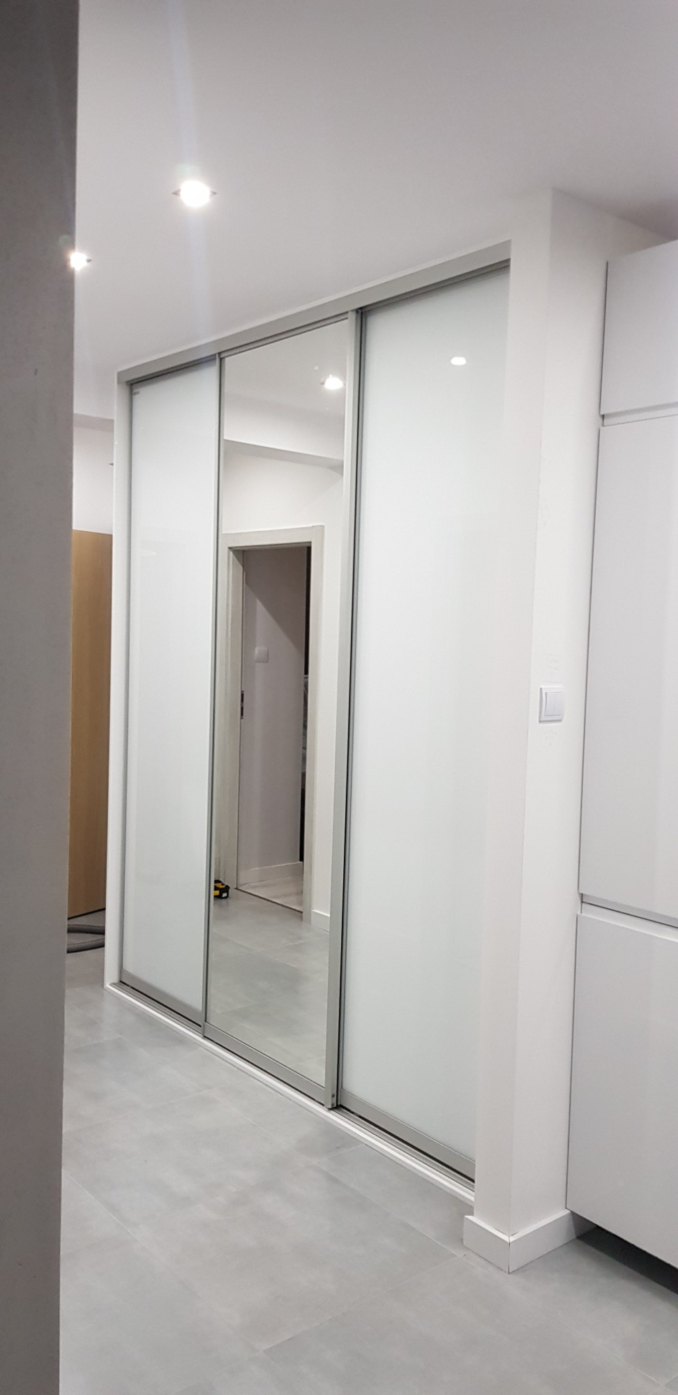 Biała szafa wnękowa z przesuwnymi drzwiami, z jednym lustrzanym panelem, w nowoczesnym wnętrzu z szarą podłogą.