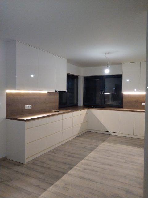 Jasna kuchnia z białymi, lakierowanymi szafkami i drewnianym blatem podświetlonym ledami, widok na dwa okna w czarnych ramach, podłoga w odcieniach szarości.