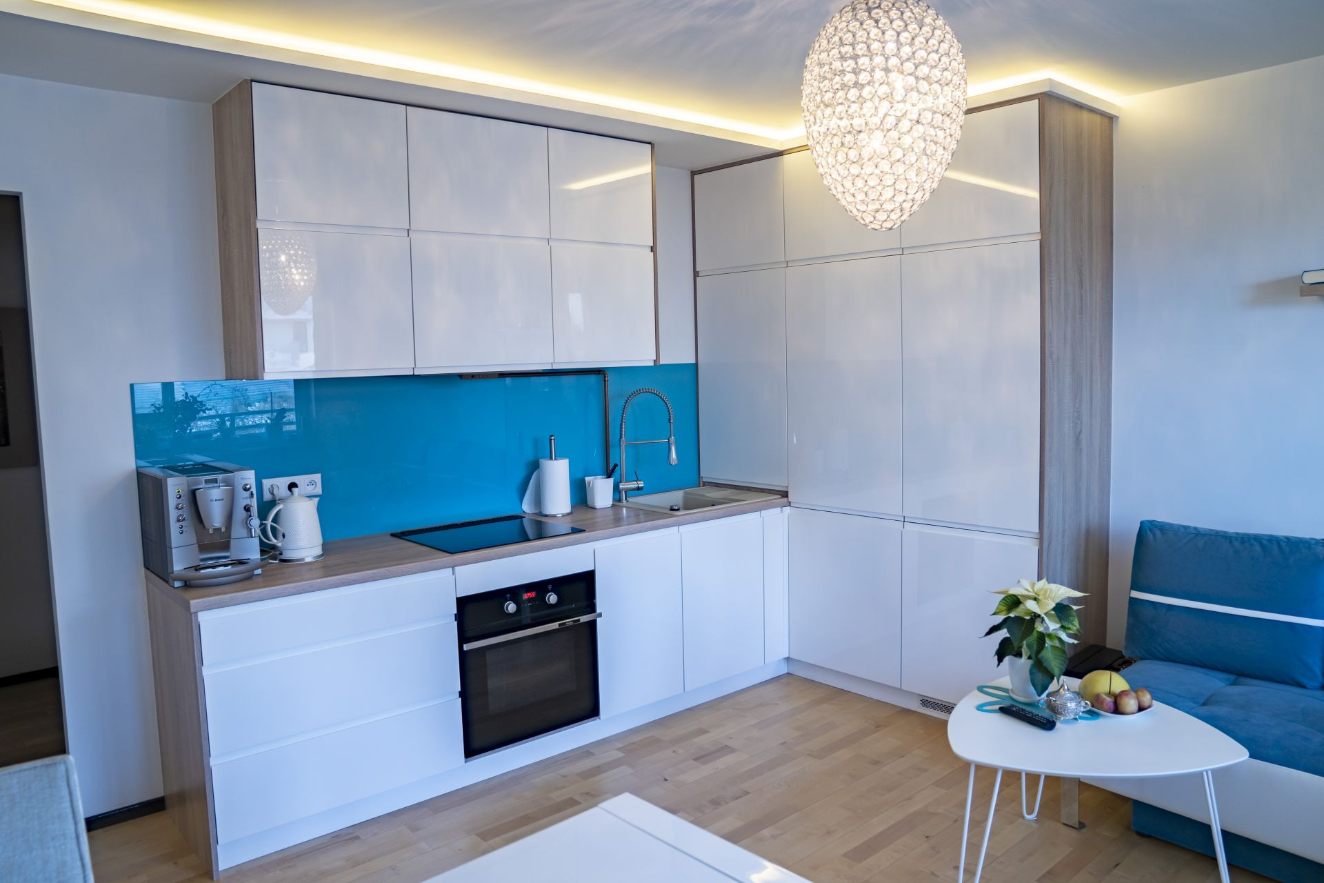Nowoczesna kuchnia z białymi, lakierowanymi frontami, drewnopodobnym wykończeniem boków i turkusowym panelem szklanym nad blatem, oświetlona wiszącą, kryształową lampą.