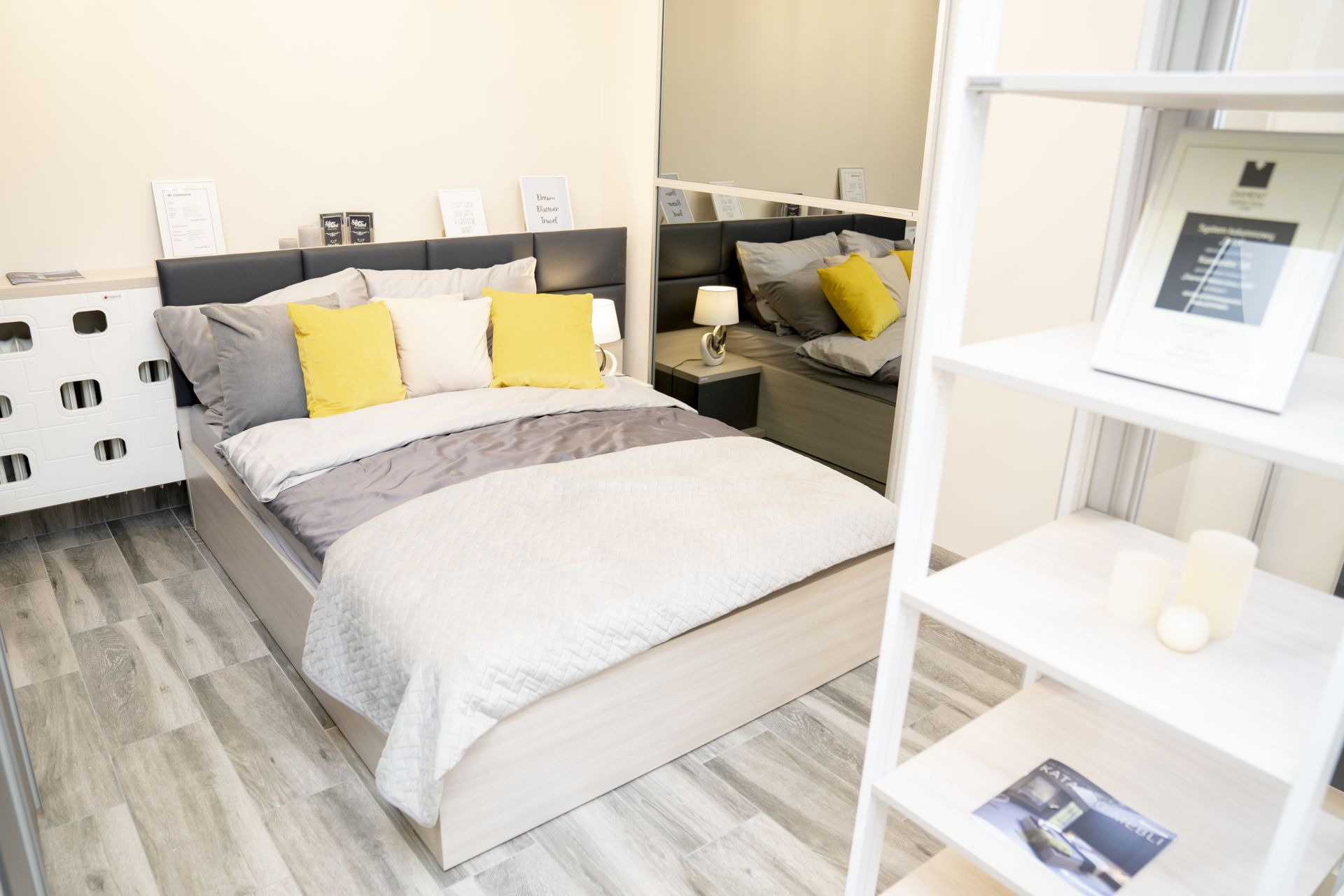 Nowoczesna sypialnia z łóżkiem kontynentalnym, szarą tapicerką zagłówka, żółtymi i szarymi poduszkami, oraz białą narzutą, widoczna szafka nocna z lampką i regał z dekoracjami.