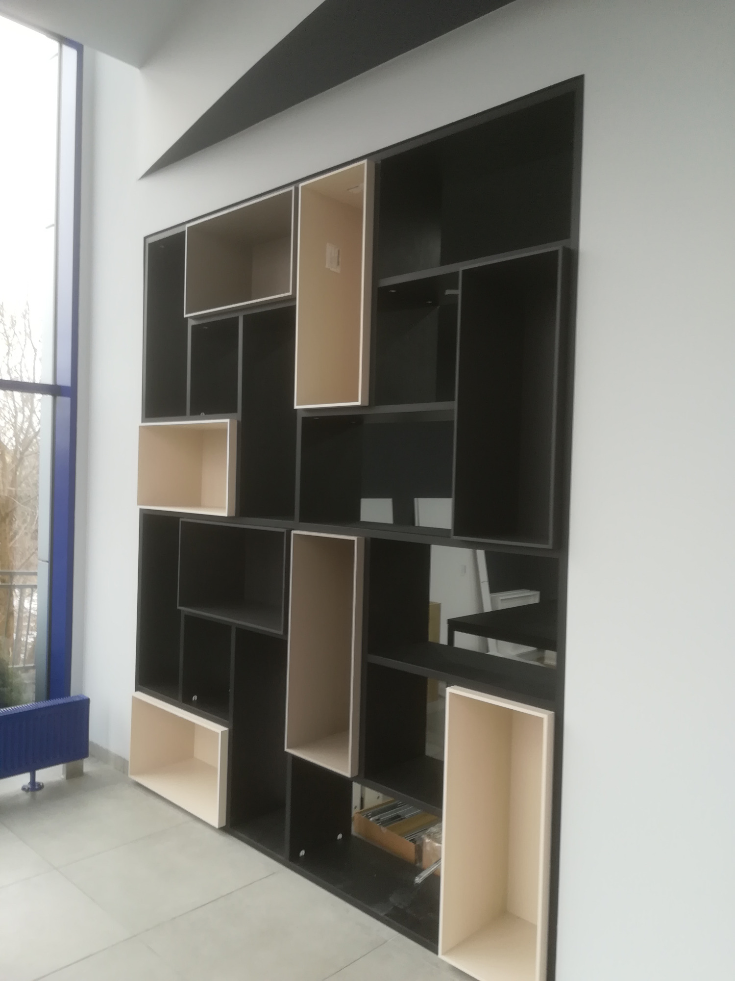 Czarna, asymetryczna szafa wnękowa z otwartymi półkami w odcieniach beżu i czerni, w nowoczesnym wnętrzu z dużym oknem.