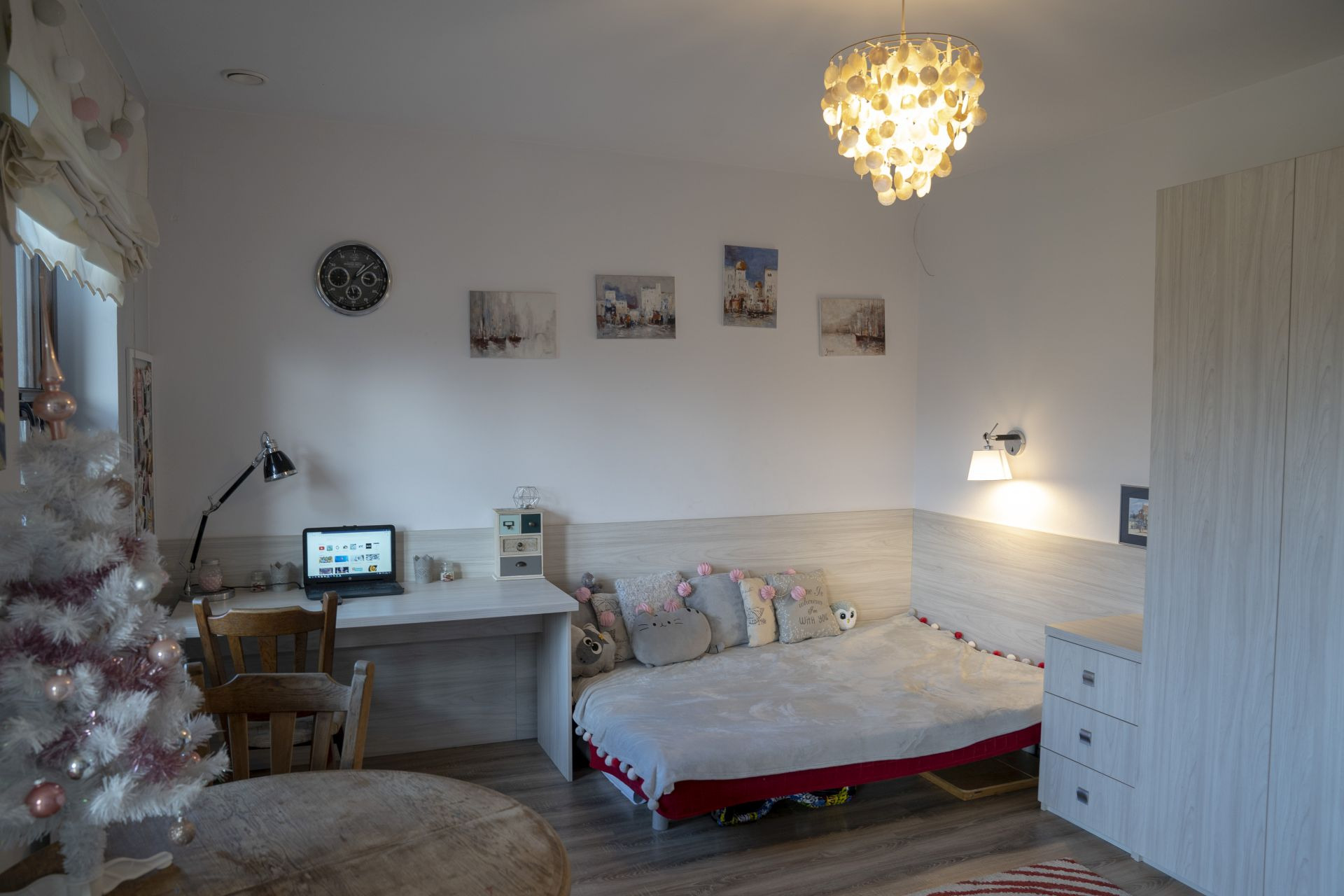 Pokój młodzieżowy z biurkiem, łóżkiem z pluszowymi poduszkami i szafą, wykończony panelami imitującymi drewno, oświetlony lampą z abażurem z muszelek.