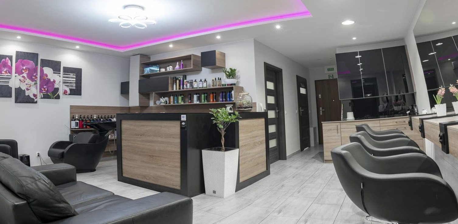 Wnętrze salonu fryzjerskiego z recepcją wykończoną drewnopodobną okleiną, czarnymi meblami i półkami z kosmetykami, oświetlone ledowym światłem w kolorze różowym.