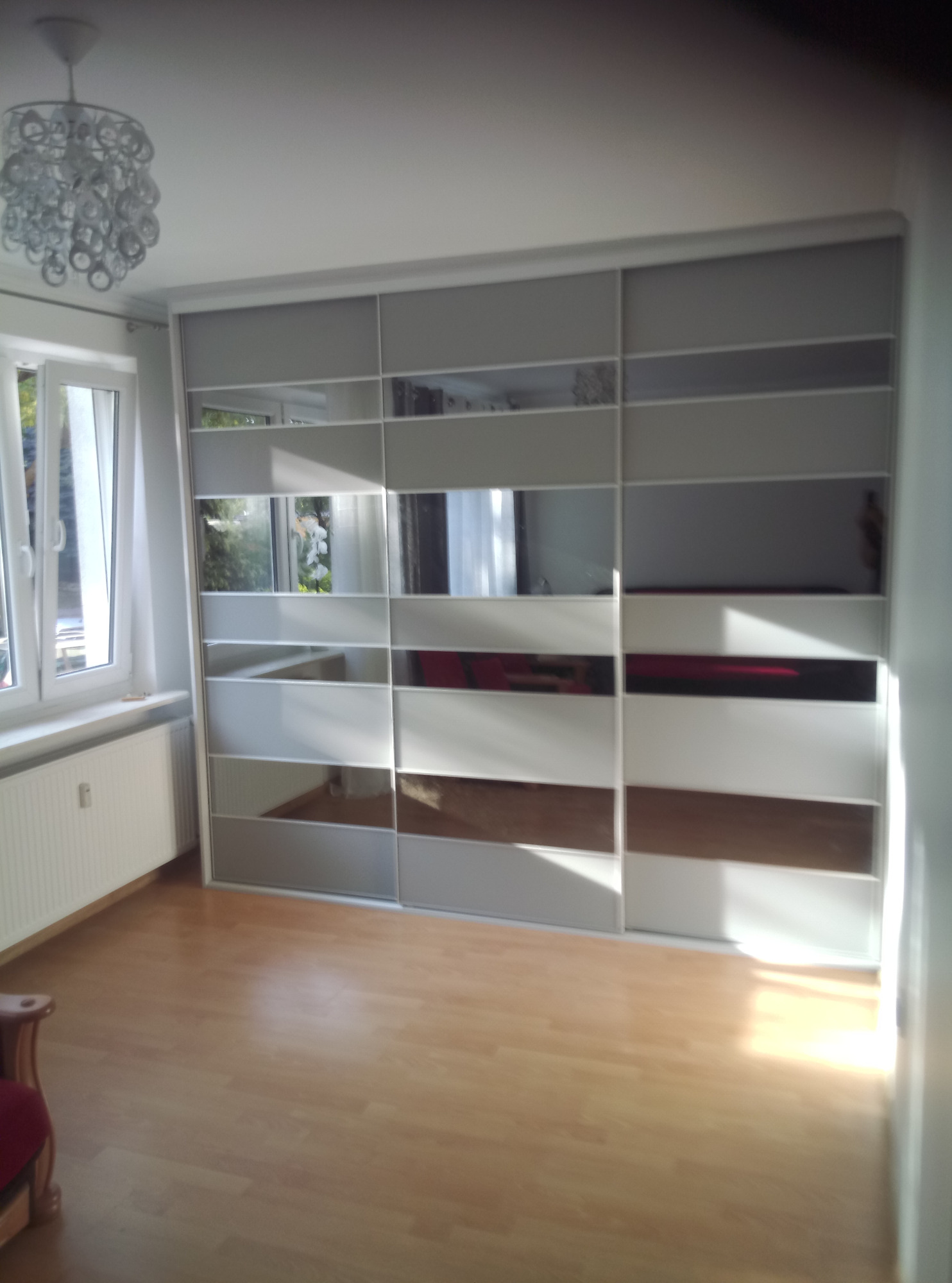 Biała szafa przesuwna z lustrzanymi panelami w pustym pokoju z parkietem, widoczny fragment krzesła z czerwonym obiciem, oświetlenie sufitowe w tle.